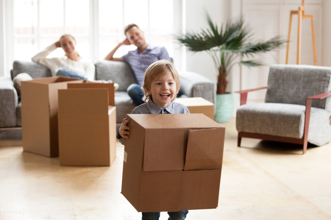 A young boy holding a cardboard box stands in front of a family about to enter their new home.